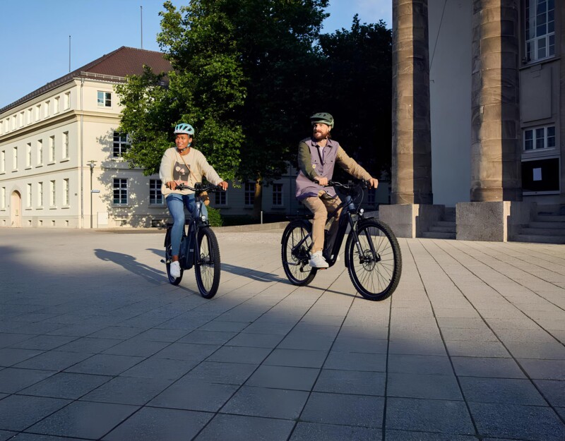 Two people are riding bikes in sunny weather in an open space. Both are wearing helmets; one person is wearing jeans and a beige sweatshirt, the other is wearing light-colored pants and a purple jacket. A building with many windows and columns is visible in the background.