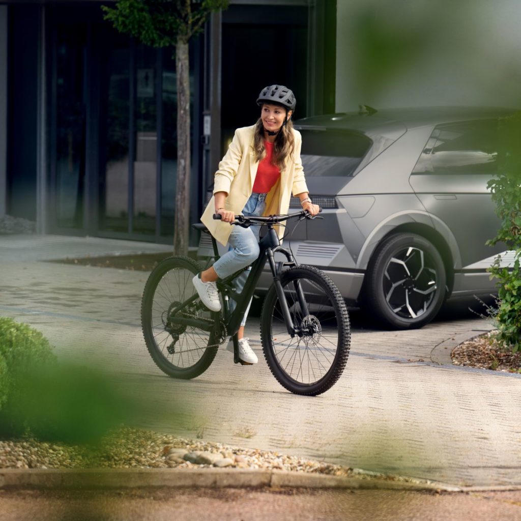 A woman wearing a helmet sits smiling on a bike, surrounded by a modern urban environment with plants in the foreground.